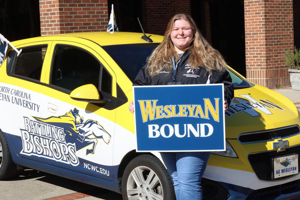 White female student holding "Wesleyan Bound" sign and posing in front of Battling Bishops car