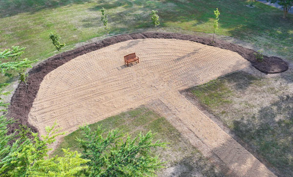 An outdoor setting with a bench on gravel surrounded by seven trees in a half moon.